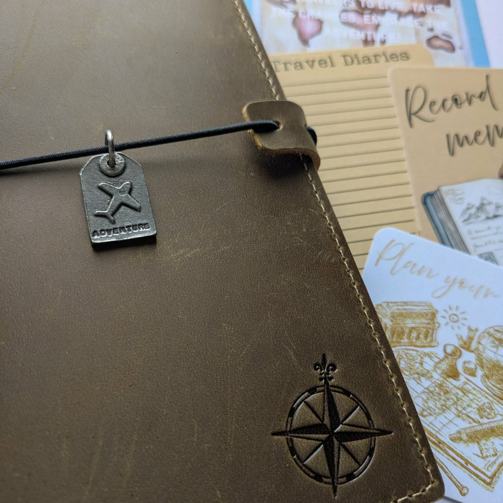 Brown leather travel diary with compass design and 'adventurer' tag, surrounded by travel-themed cards.