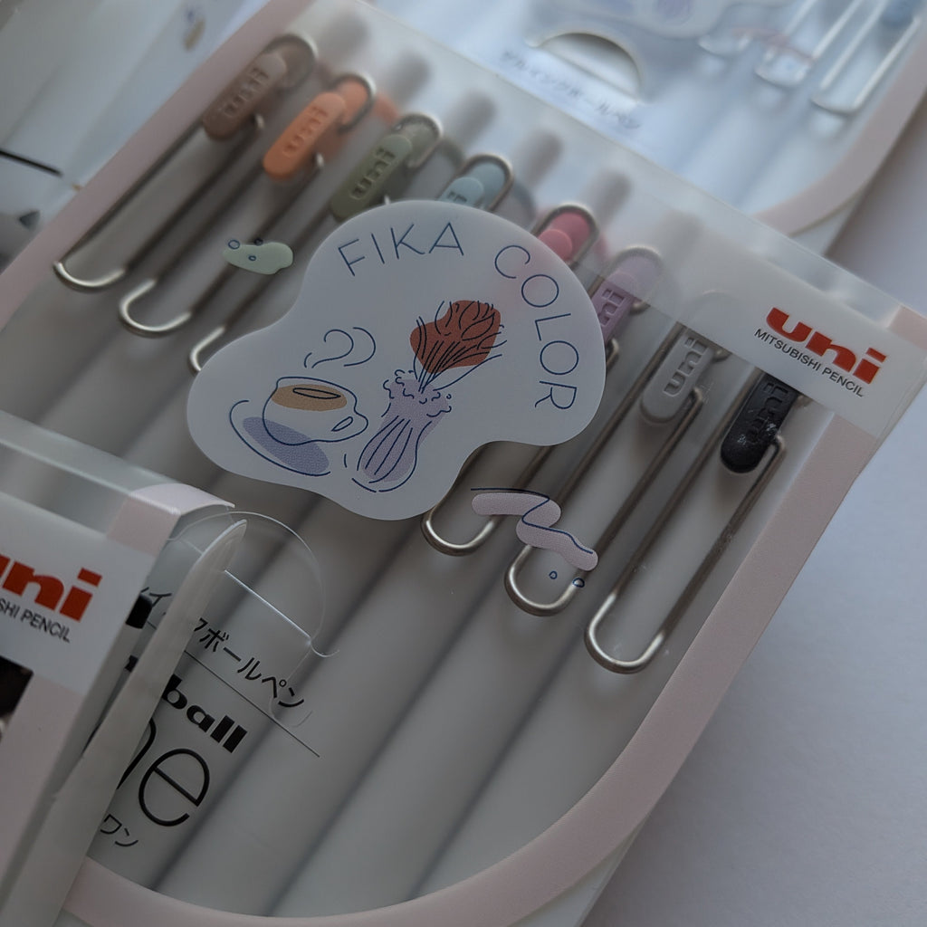 Uni ball pen accessories set in a clear plastic case, featuring silver metal clips with pastel-colored tips and a decorative sticker that reads 'FIKA COLOR'.
