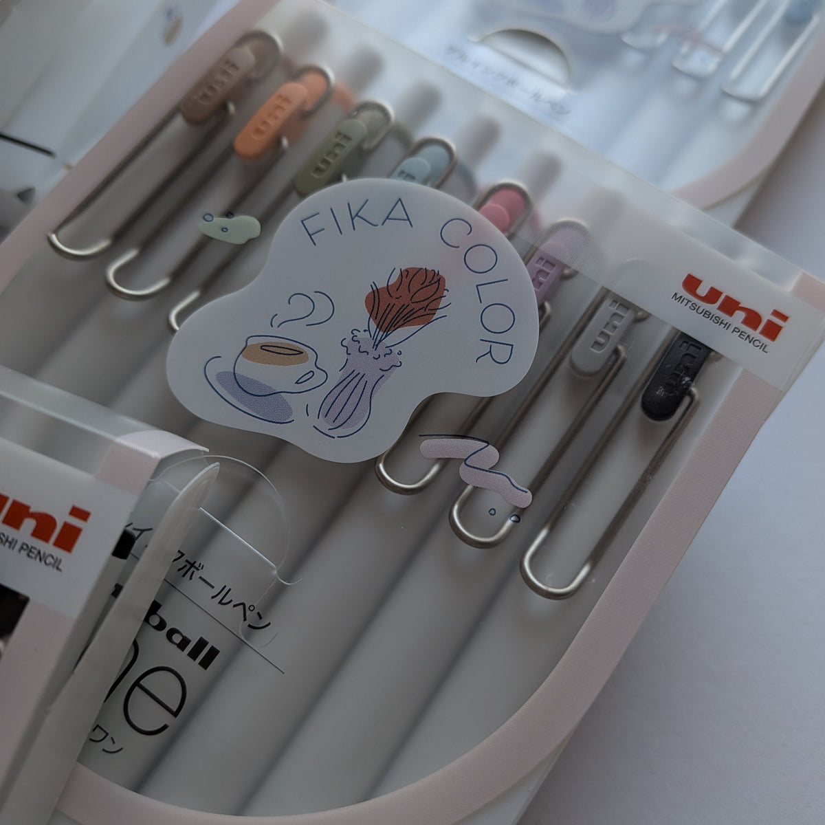 Uni ball pen accessories set in a clear plastic case, featuring silver metal clips with pastel-colored tips and a decorative sticker that reads 'FIKA COLOR'.
