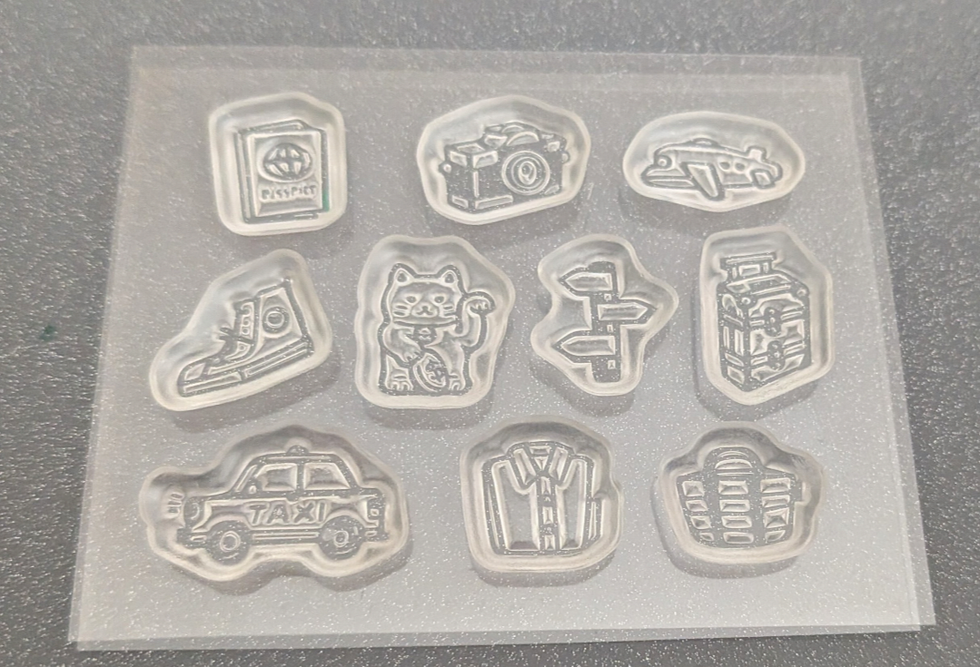 A clear silicone stanp with 10 stamps featuring different illustrated designs, including a passport, camera, airplane, sneaker, cat, road signs, bottles, a taxi, a jacket, and headphones, photographed on a gray textured surface.