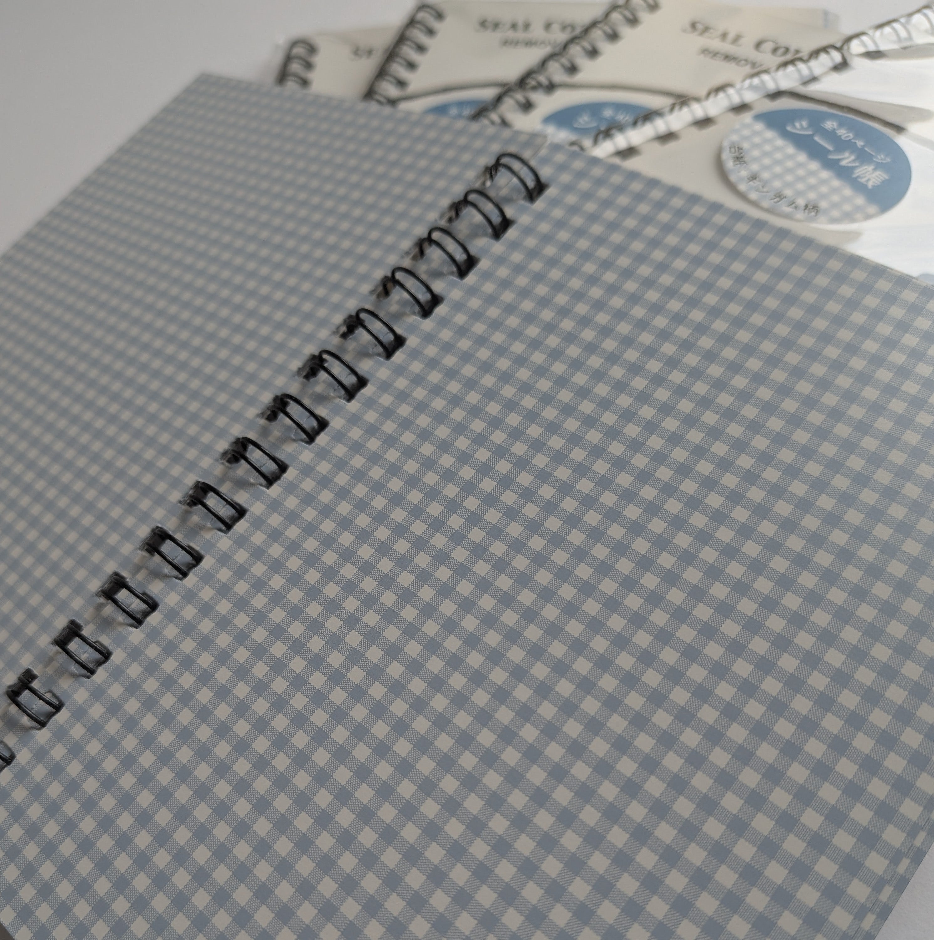 A close-up of a spiral-bound notebook or journal with a light blue and white gingham cover, with additional similar notebooks partially visible behind it on a plain light background.