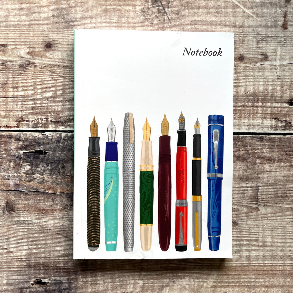 A white notebook with the word 'Notebook' printed at the top right, featuring an illustration of eight fountain pens in various colors and styles, displayed on a rustic wooden surface.