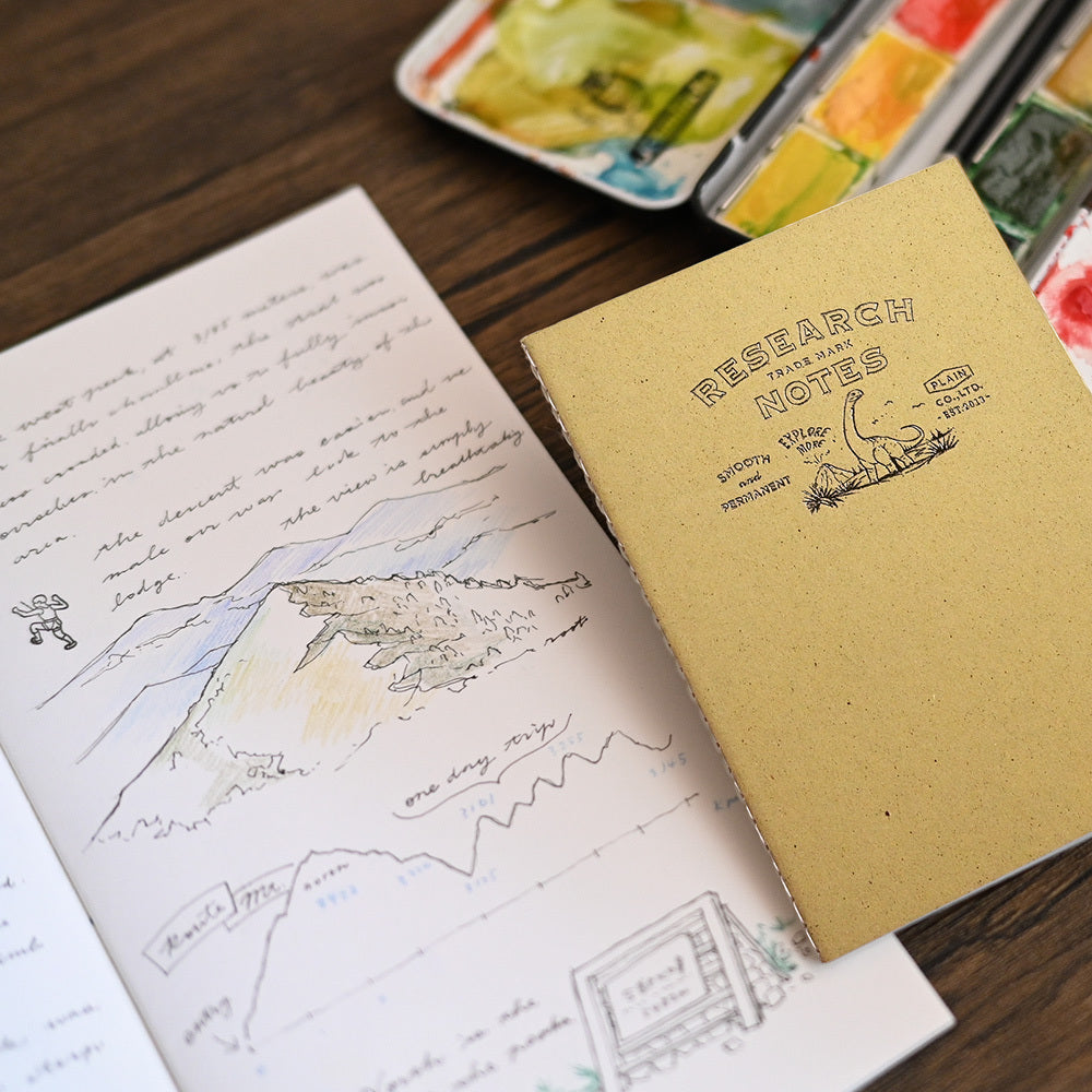 A paper notebook labeled 'RESEARCH NOTEBOOK' lies on a wooden table next to an open sketchbook with handwritten notes and a landscape drawing. Watercolor paint palettes are partially visible in the background.