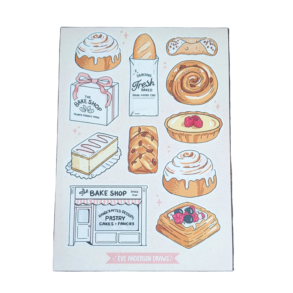 A rectangular printed card featuring illustrated baked goods and a bakery storefront, placed on a dark wood surface. The card includes drawings of pastries, breads, tarts, and a bakery shop with small pink decorative accents.