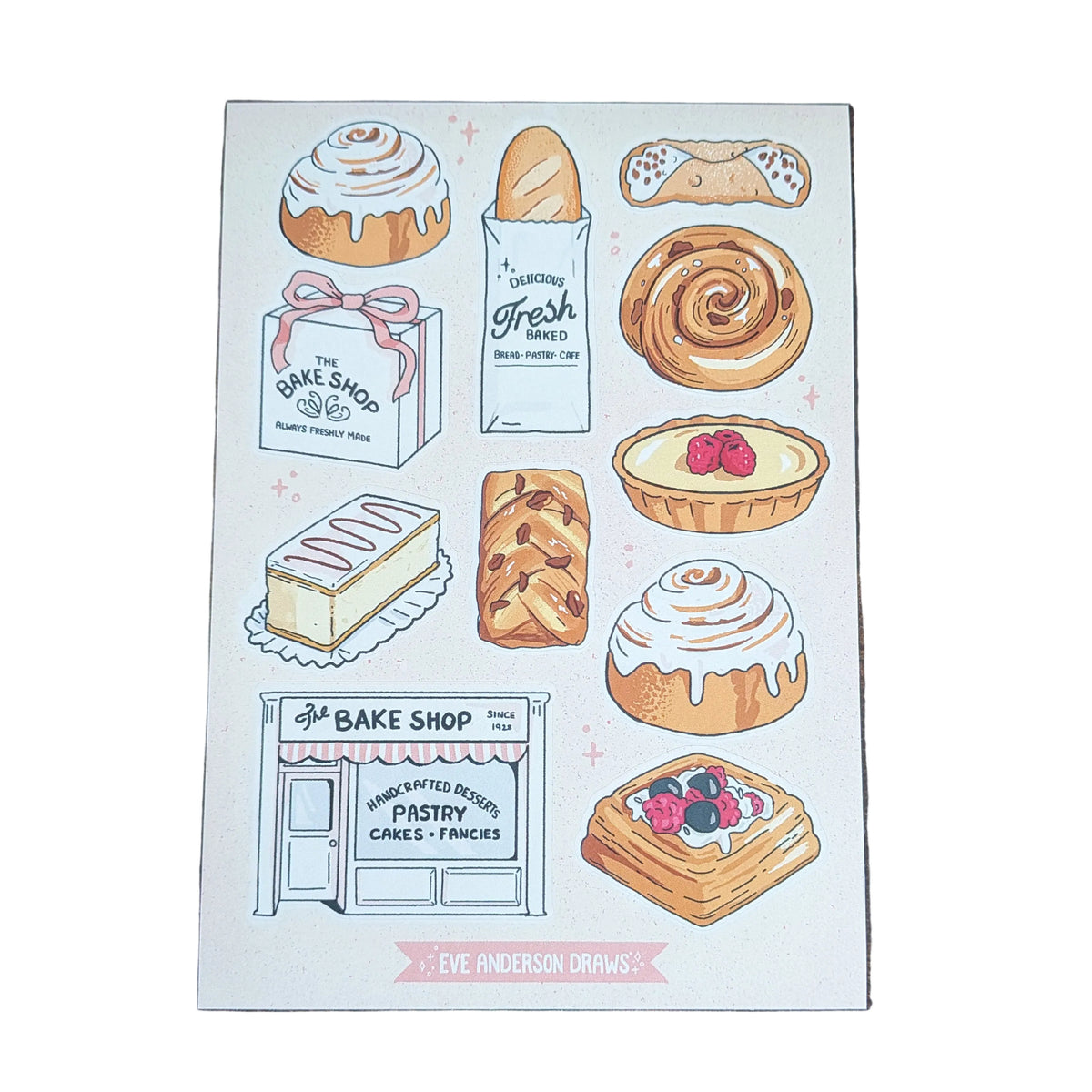 A rectangular printed card featuring illustrated baked goods and a bakery storefront, placed on a dark wood surface. The card includes drawings of pastries, breads, tarts, and a bakery shop with small pink decorative accents.