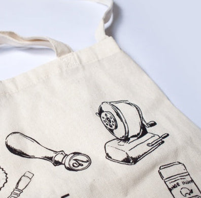 A close-up of a cream canvas tote bag with black line-art illustrations of sewing tools, including a rotary cutter, shown on a white background.