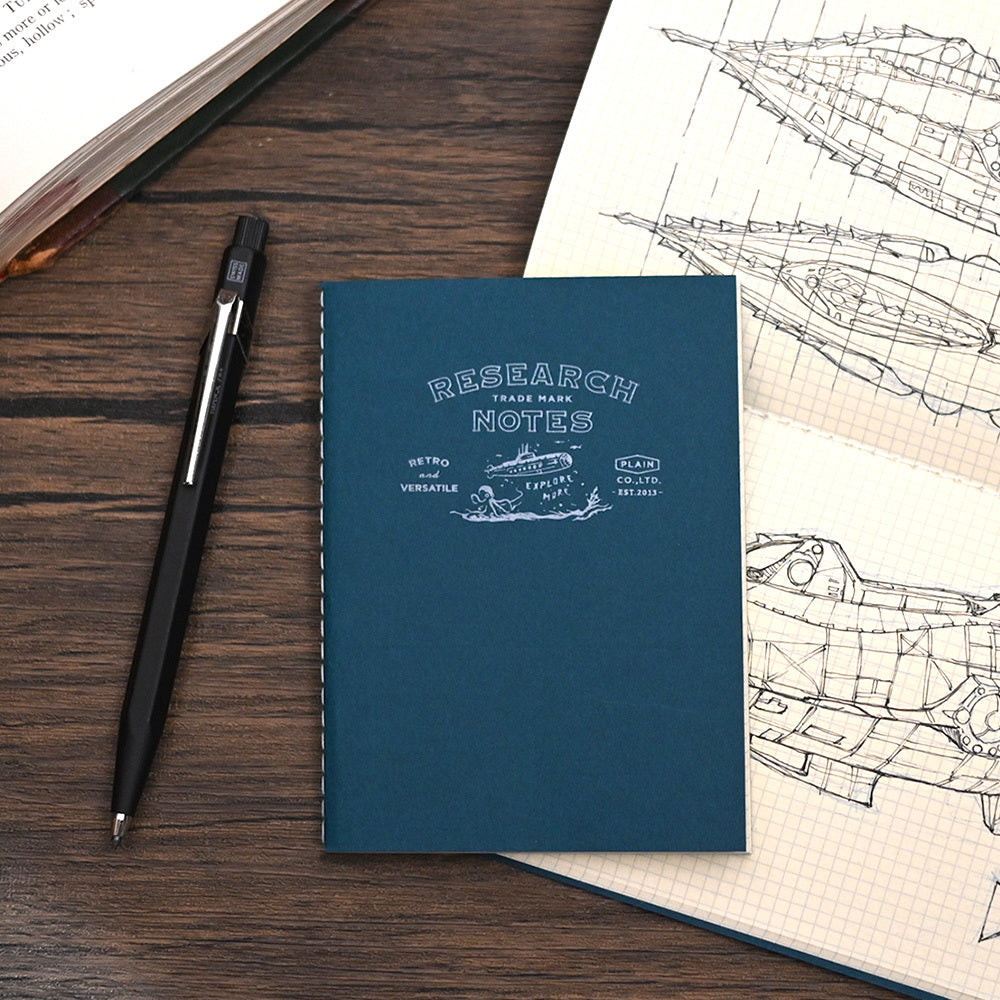A dark teal blue spiral-bound notebook with white printed text reading 'RESEARCH NOTES' is placed on a dark wood desk next to a black pen and an open sketchbook with technical drawings.