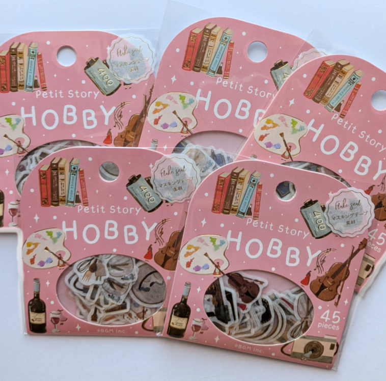 Four pink retail packages of stickers featuring vintage illustrations of books, bottles, and violins, with a clear window showing the included designs. The packages are labeled 'Petit Story HOBBY' and '45 pieces'.