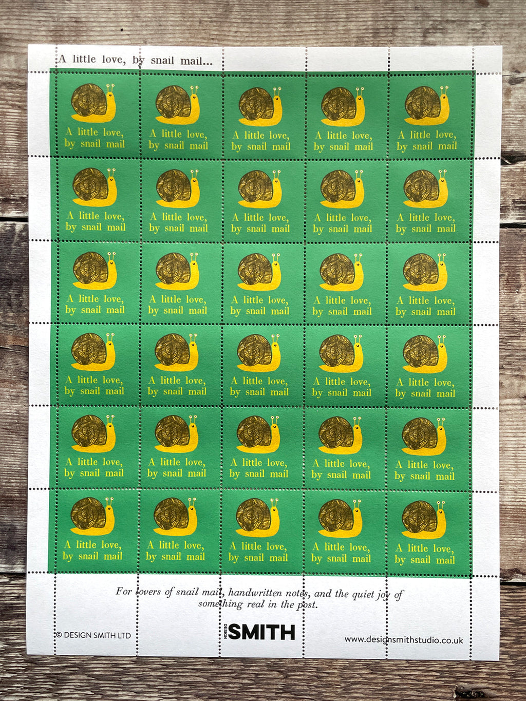 A sheet of postage stamps laid flat on a rustic wooden surface. Each stamp has a green background with a yellow crescent and brown snail illustration, plus the text 'A little love, by small mail.
