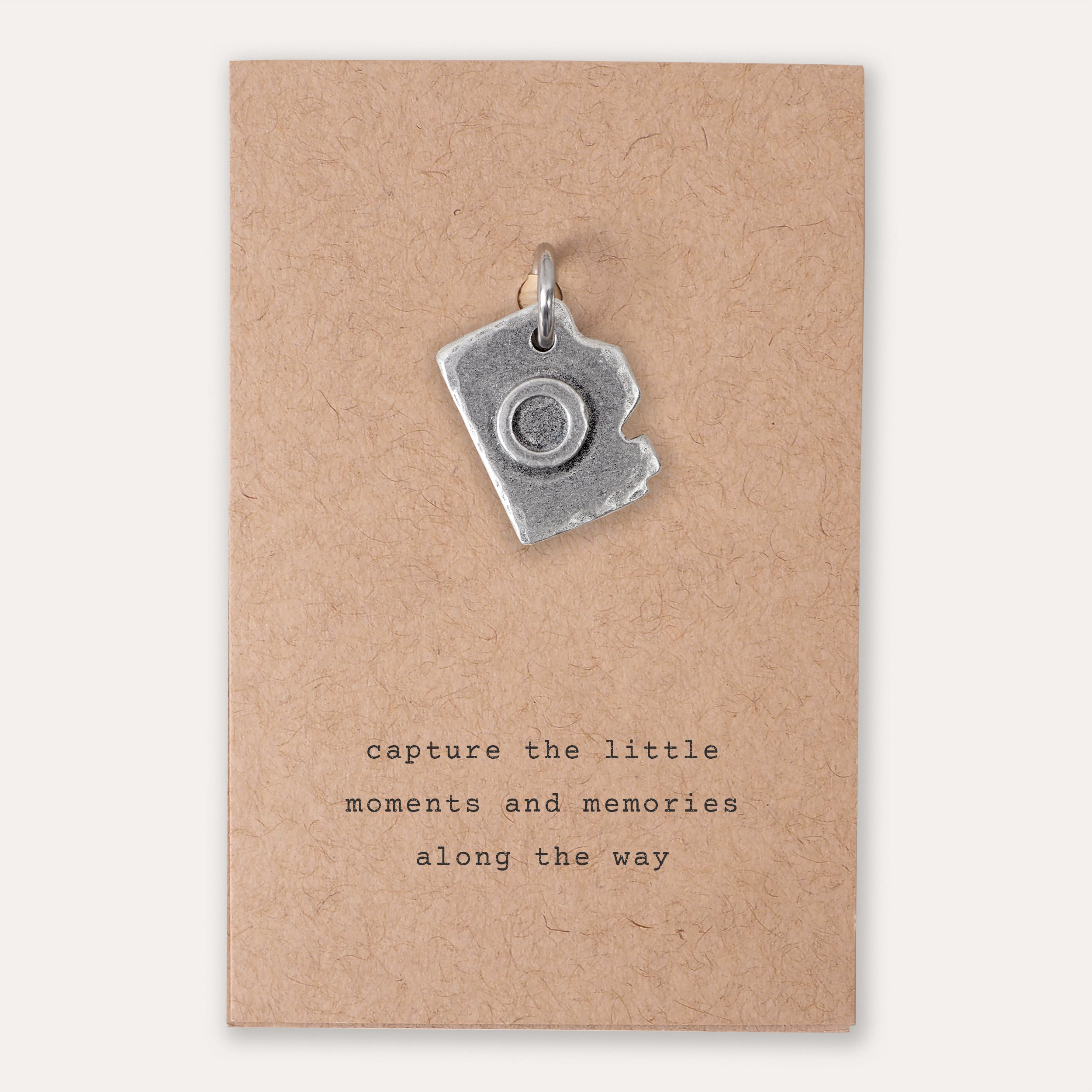 A small silver camera-shaped charm on a kraft paper card with the printed phrase 'capture the little moments and memories along the way'.