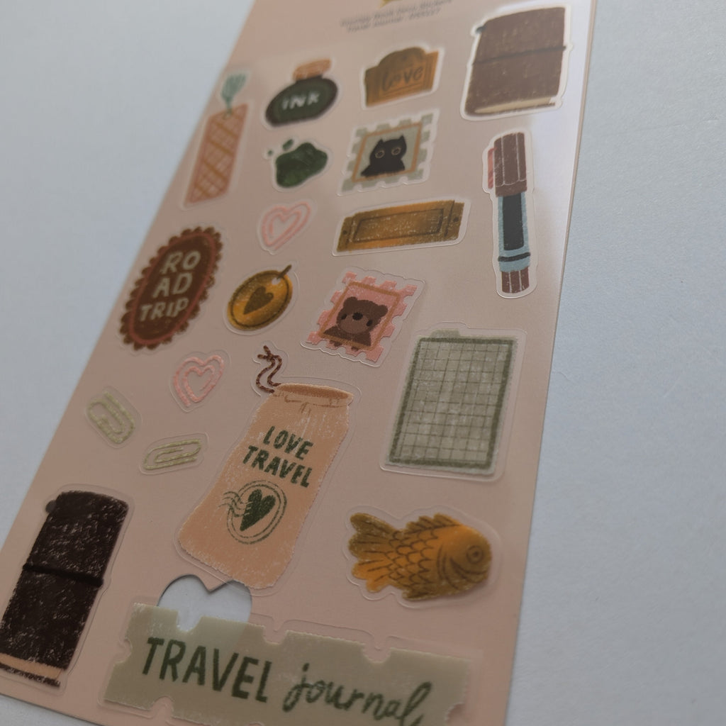 A sheet of travel-themed stickers on a light peach backing card, featuring retro stamps, a suitcase, a fish, a book, and phrases like 'ROAD TRIP', 'LOVE TRAVEL', and 'TRAVEL journal' on a plain light gray background.