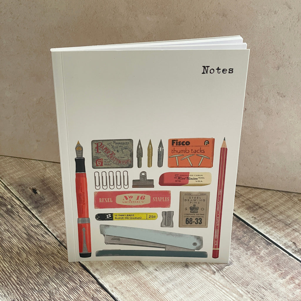 A white hardcover notebook standing upright on a rustic wooden surface against a textured beige wall. The cover features a collage of vintage stationery items, including a red fountain pen, pencils, stamps, clips, and a stapler, with the word 'Notes' printed in black at the top right.
