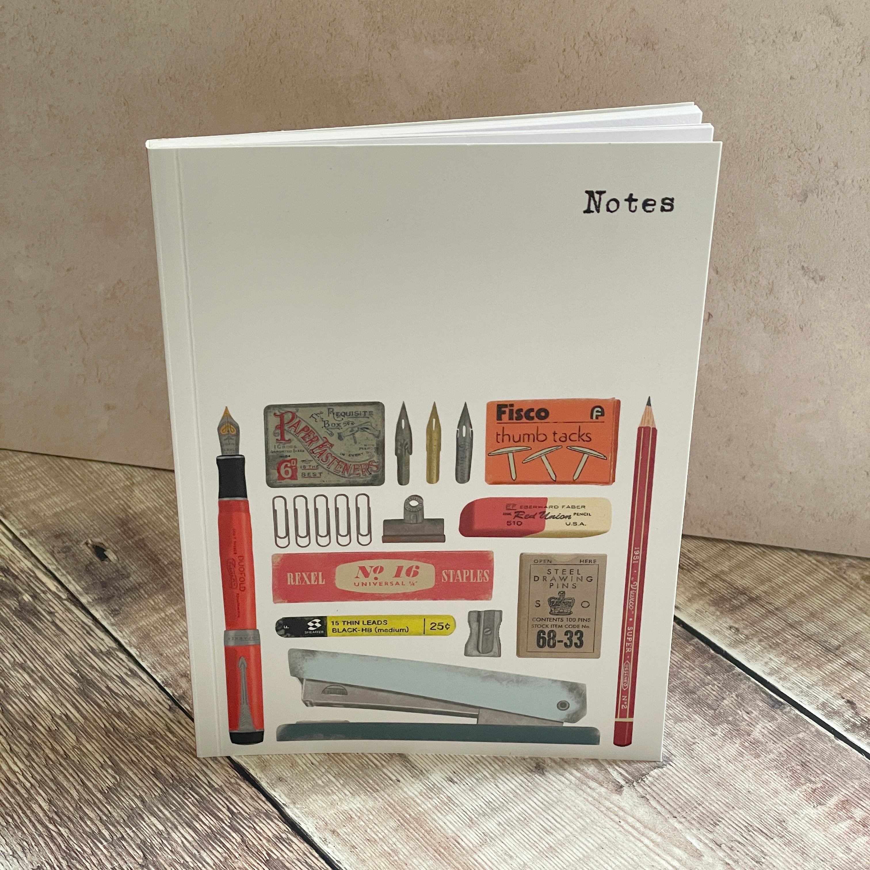 A white hardcover notebook standing upright on a rustic wooden surface against a textured beige wall. The cover features a collage of vintage stationery items, including a red fountain pen, pencils, stamps, clips, and a stapler, with the word 'Notes' printed in black at the top right.