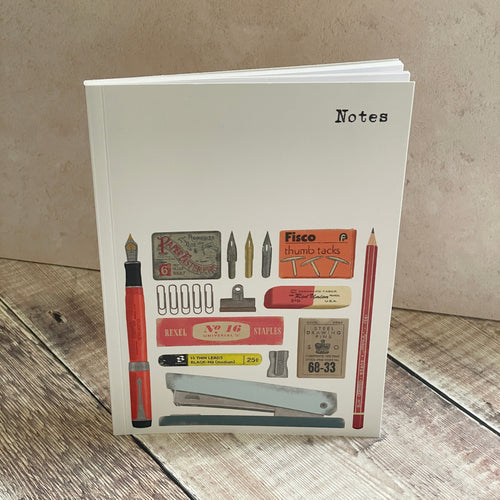 A white hardcover notebook standing upright on a rustic wooden surface against a textured beige wall. The cover features a collage of vintage stationery items, including a red fountain pen, pencils, stamps, clips, and a stapler, with the word 'Notes' printed in black at the top right.