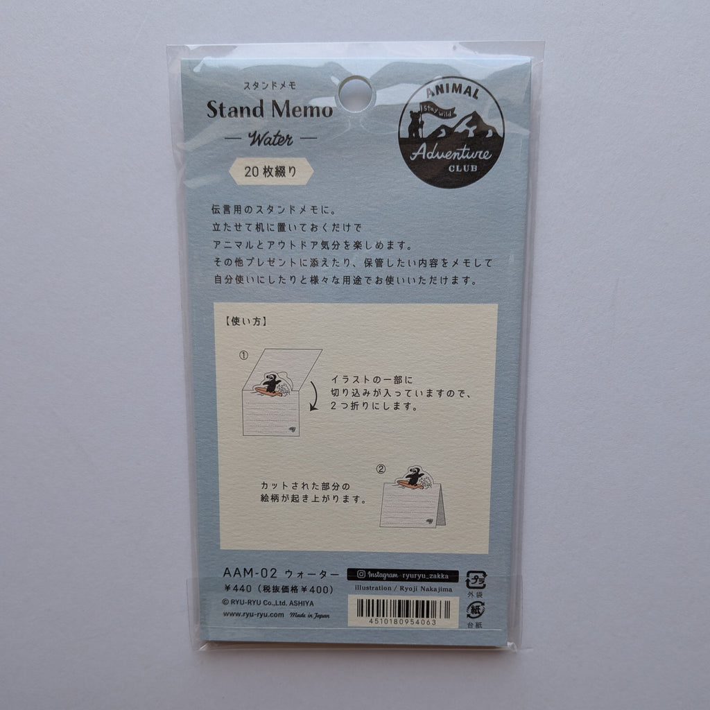 The back of a packaged Japanese stand memo pad, sealed in a clear plastic sleeve and photographed against a plain light background. The card is pale blue-gray with instructional diagrams, product text, and a circular logo reading Animal Adventures.
