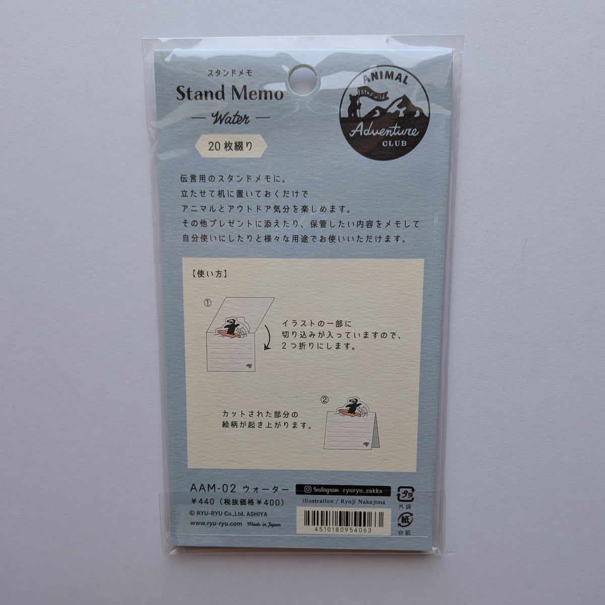 The back of a packaged Japanese stand memo pad, sealed in a clear plastic sleeve and photographed against a plain light background. The card is pale blue-gray with instructional diagrams, product text, and a circular logo reading Animal Adventures.