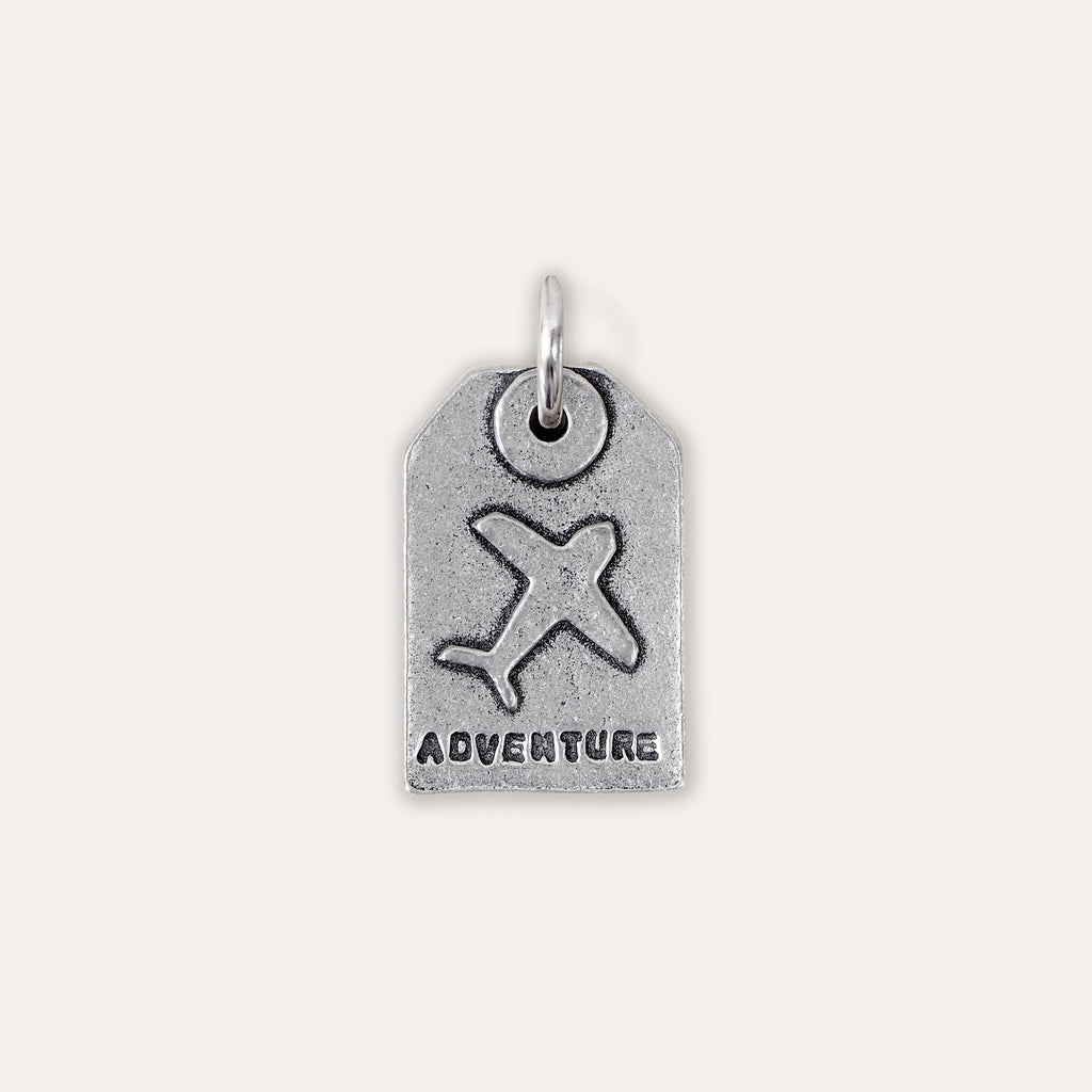 A silver-toned luggage tag charm featuring an embossed airplane and the word 'ADVENTURE' at the bottom, shown on a plain light beige background.