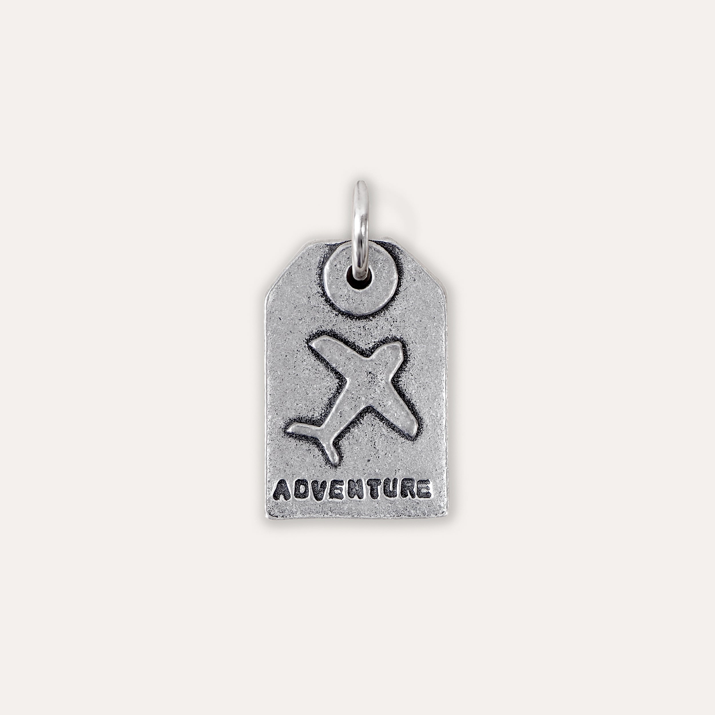 A silver-toned luggage tag charm featuring an embossed airplane and the word 'ADVENTURE' at the bottom, shown on a plain light beige background.