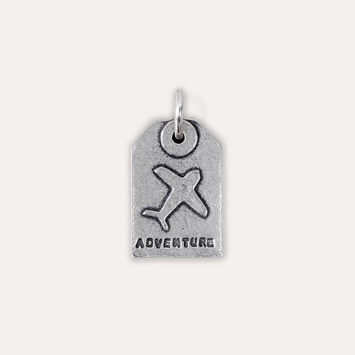 A silver-toned luggage tag charm featuring an embossed airplane and the word 'ADVENTURE' at the bottom, shown on a plain light beige background.
