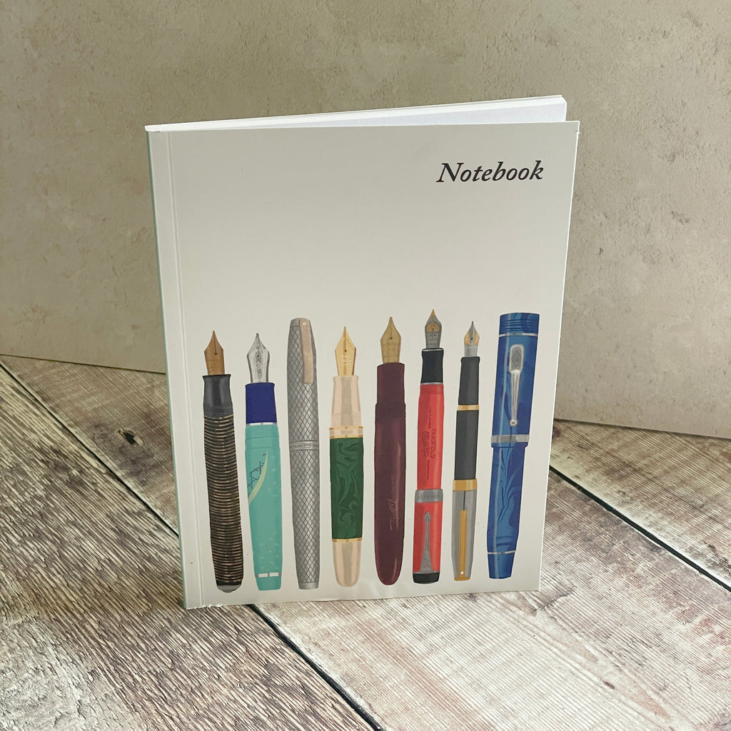 A notebook with a white cover featuring an illustration of eight vintage fountain pens, standing on a rustic wooden surface against a textured beige wall.