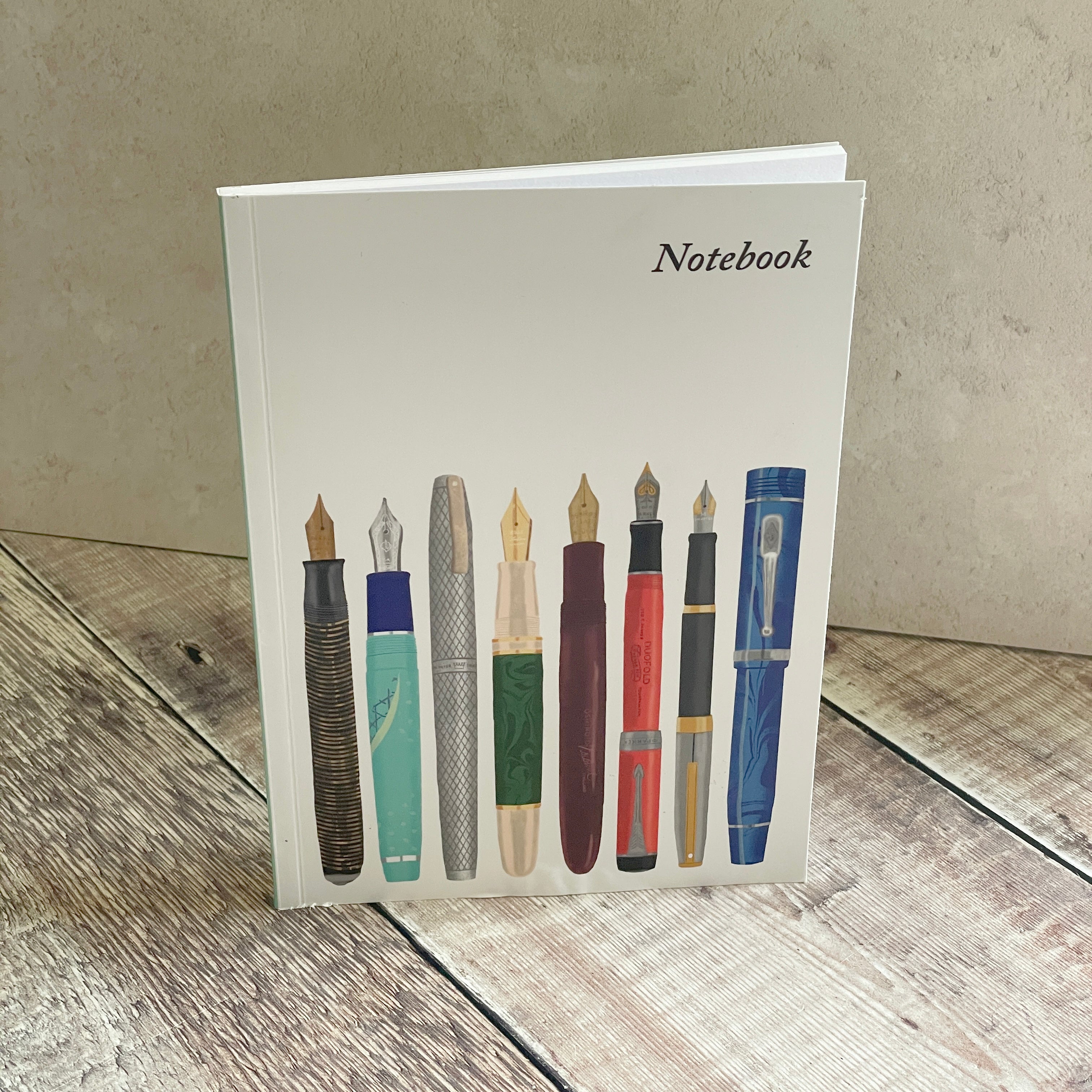 A notebook with a white cover featuring an illustration of eight vintage fountain pens, standing on a rustic wooden surface against a textured beige wall.