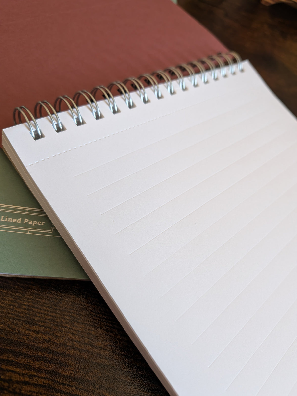 An open spiral-bound lined notebook lying on a dark wooden surface, with a muted green book or notebook underneath and a dusty rose cover visible behind it.