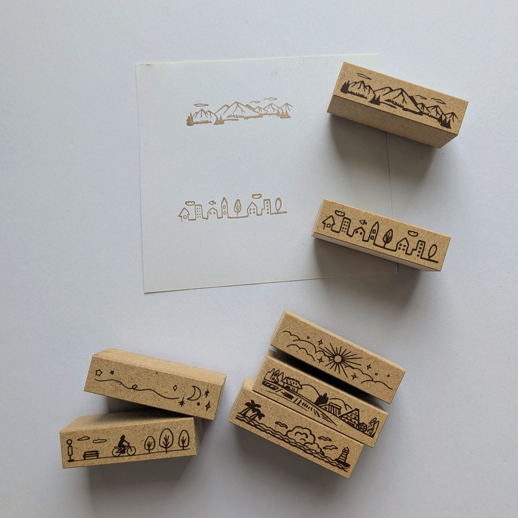 A set of wooden rubber stamps with engraved landscape and travel-themed illustrations, arranged on a white surface next to a white card that shows the stamped designs.