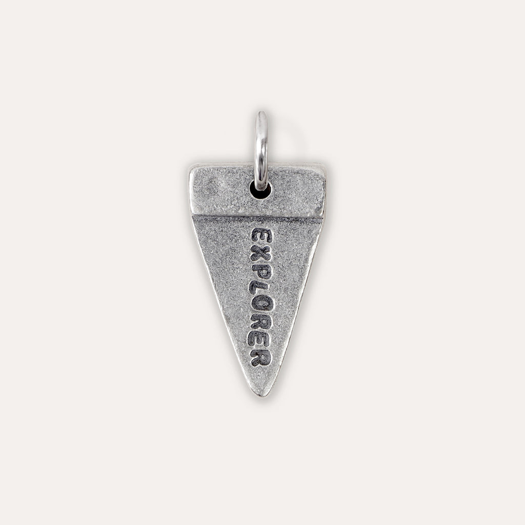 A silver triangular pendant charm engraved with the word 'EXPLORER' vertically, shown on a light beige background.