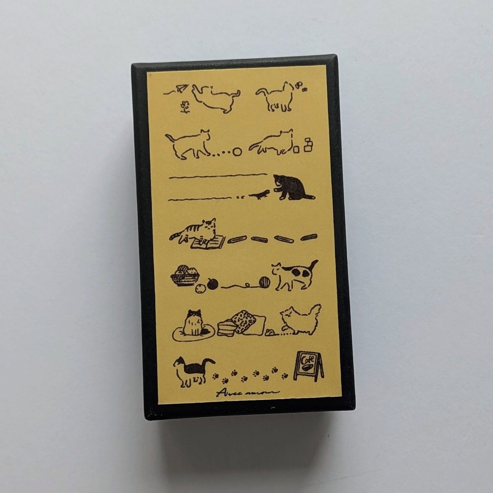 A rectangular black box with a yellow front panel featuring black line drawings of cats and cat-related illustrations, photographed on a plain light background.