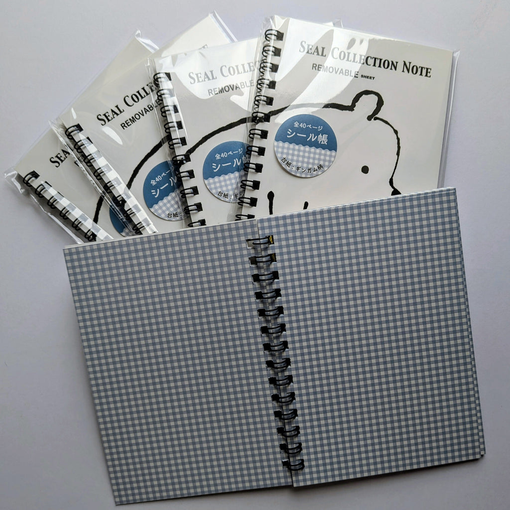 A set of spiral-bound notebook covers or note pads with a blue-gray and white gingham pattern, shown with several individually wrapped items fanned behind them on a light gray background. The front item is open to show the black spiral binding.