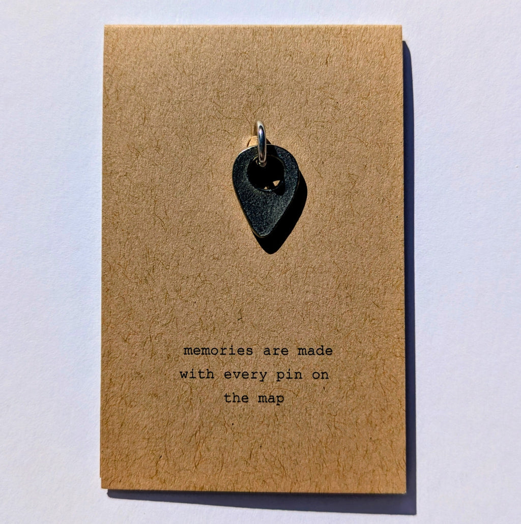 Metal map pin on a brown card with text 'memories are made with every pin on the map' on a light gray background