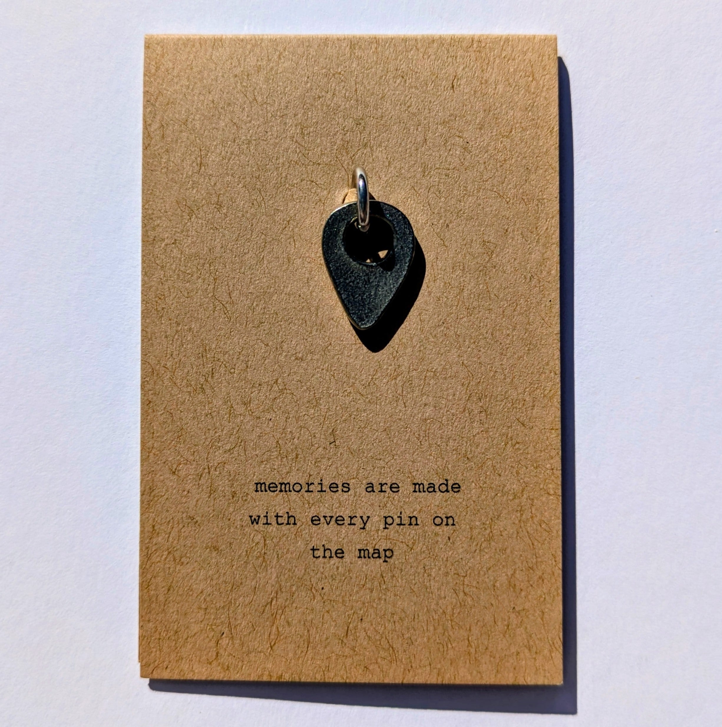 Metal map pin on a brown card with text 'memories are made with every pin on the map' on a light gray background