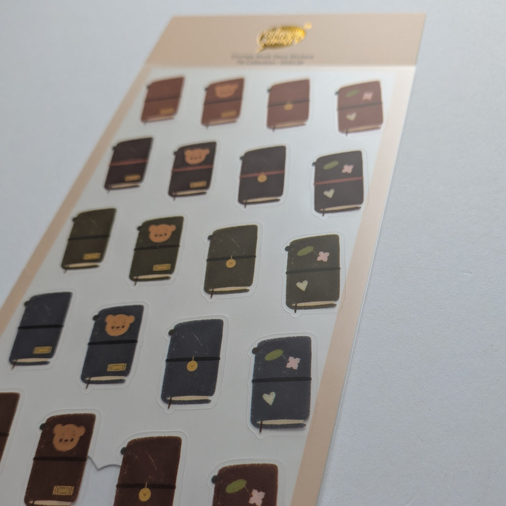 A sheet of planner stickers featuring cute illustrated notebooks in brown, black, and navy, displayed on a light gray background.