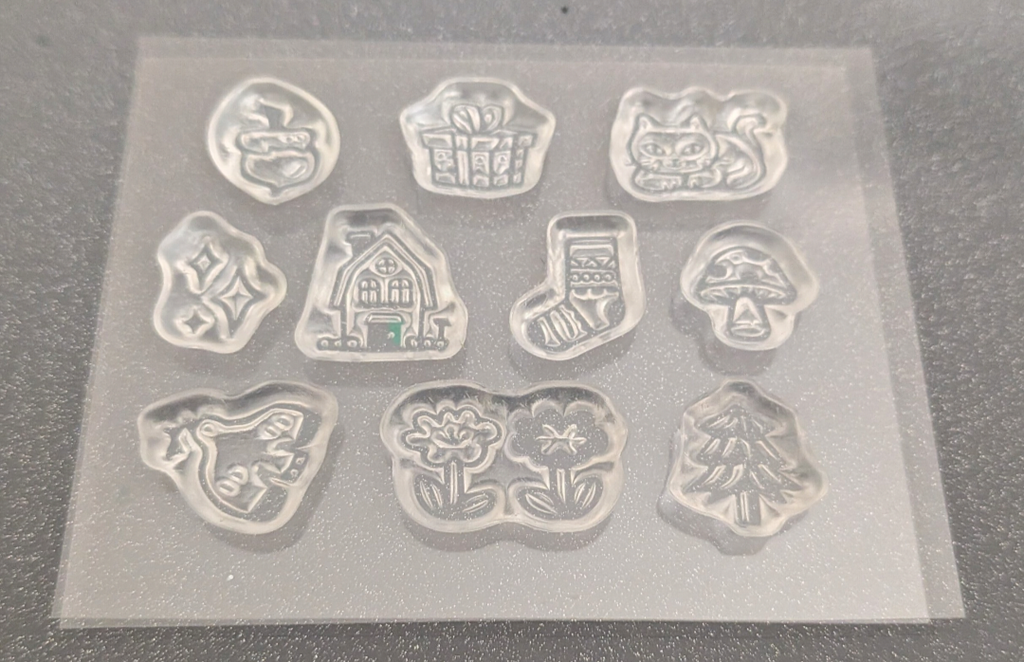 10 individual clear stamps, each shaped like a different whimsical motif including a gift box, cat, mushroom, house, stocking, and trees, shown on a dark textured surface.