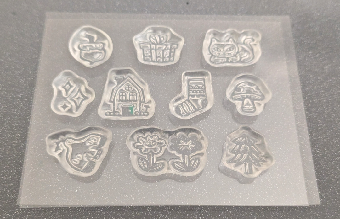 10 individual clear stamps, each shaped like a different whimsical motif including a gift box, cat, mushroom, house, stocking, and trees, shown on a dark textured surface.