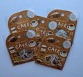 Four small retail packages of decorative stickers arranged on a light blue background. Each package is brown with coffee-themed illustrations and the text 'Petit Story CAFE' and '45 pieces'.