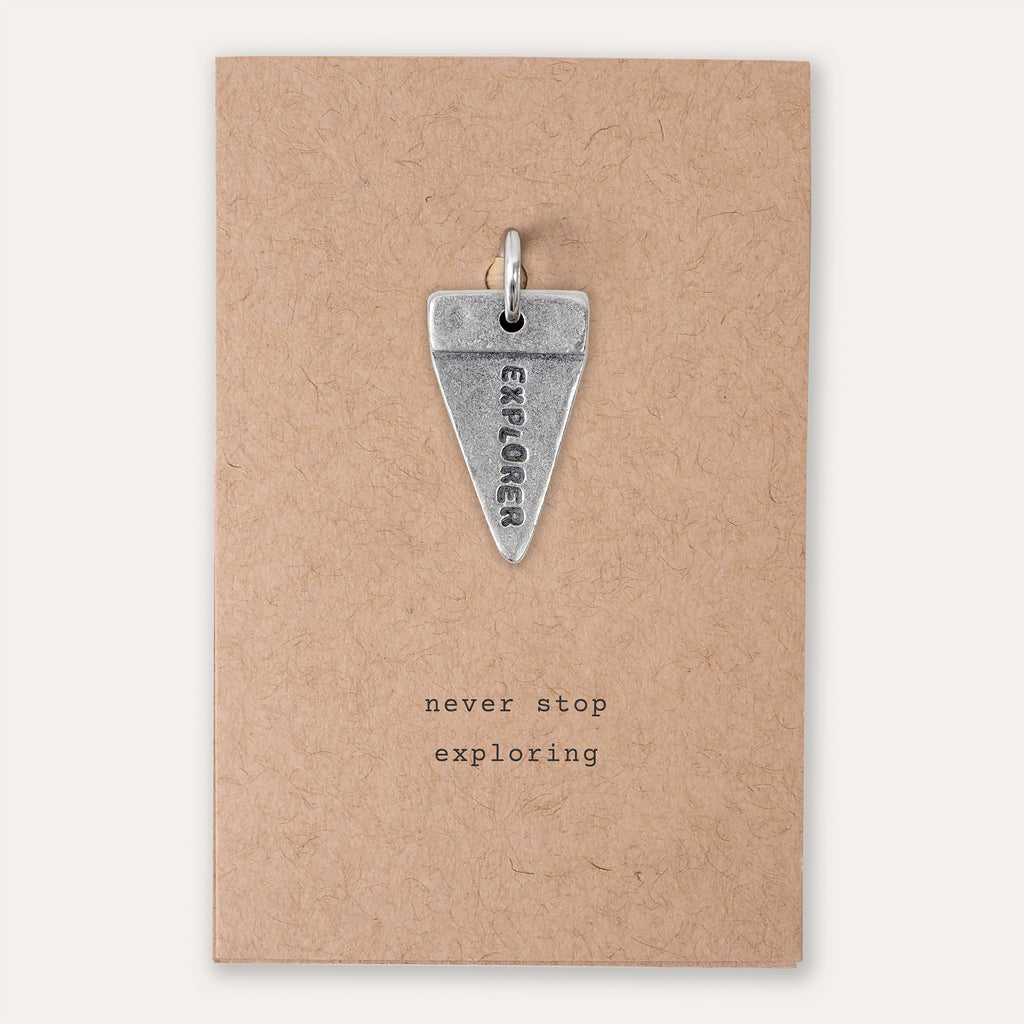 A silver triangular pendant engraved with the word 'EXPLORER' and a small loop at the top, displayed on a kraft paper backing card with the printed phrase 'never stop exploring'.