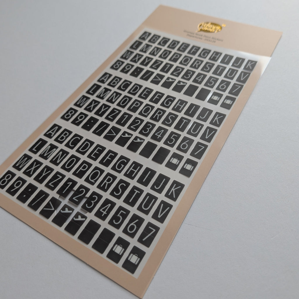 A sheet of black alphabet and number stickers on a beige backing card, photographed at an angle on a light gray background.