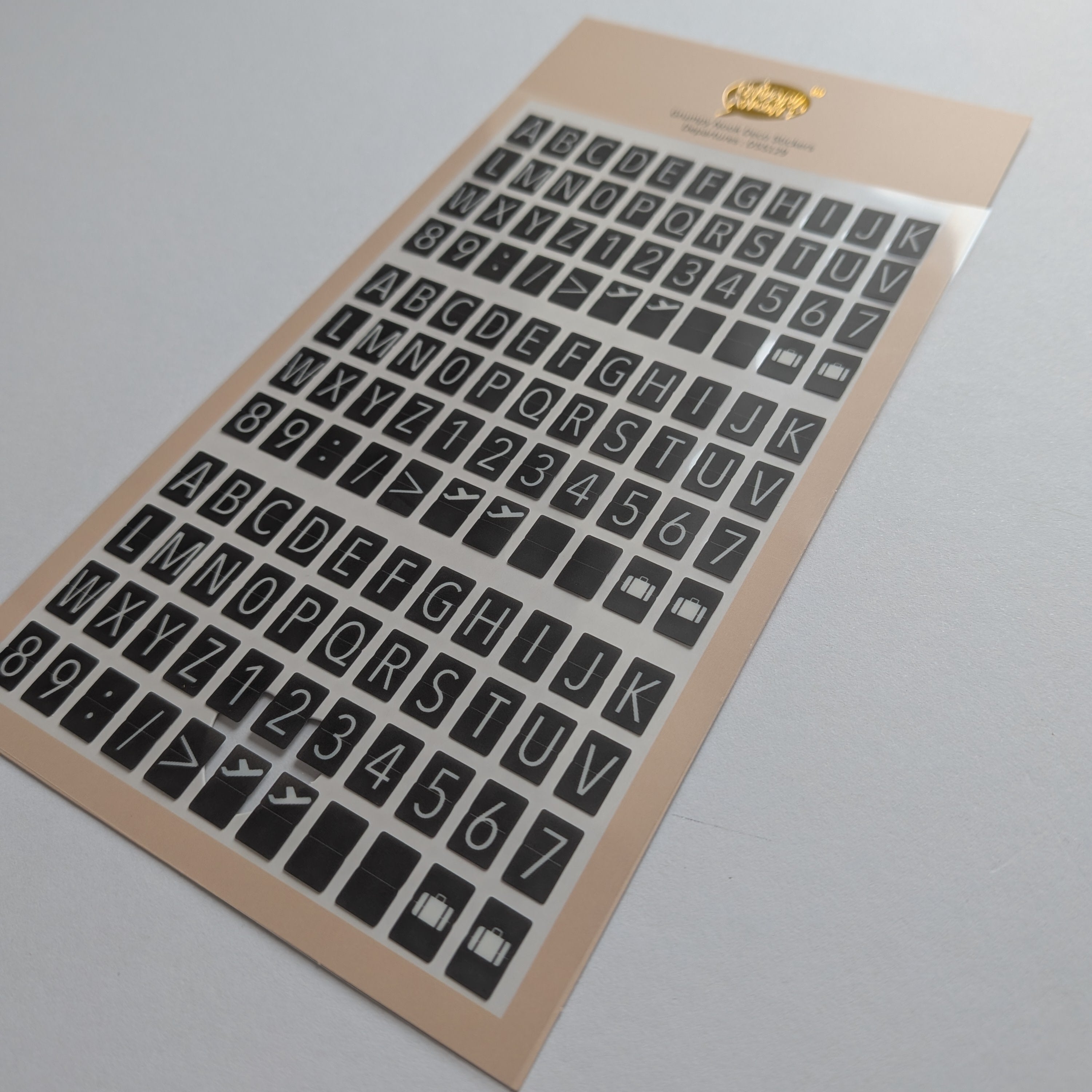 A sheet of black alphabet and number stickers on a beige backing card, photographed at an angle on a light gray background.