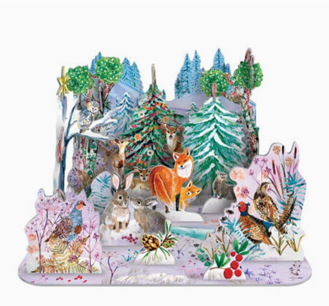 A colorful 3D pop-up cardboard forest scene featuring woodland animals such as foxes, rabbits, deer, a owl, and birds among decorated Christmas trees, flowers, and snowy ground on a white background.