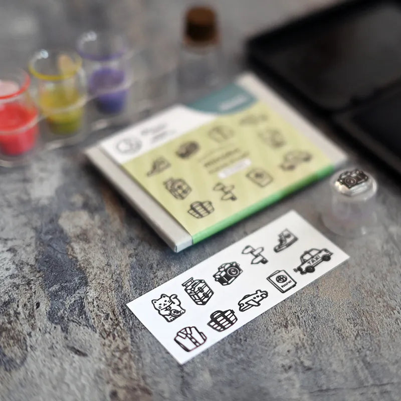 A sheet of small black outline icons or stamps placed on a gray stone-like surface, with a folded booklet or card behind it and a small clear container with a round object nearby. The scene appears styled in a craft or stationery setting, but no person or animal is present.