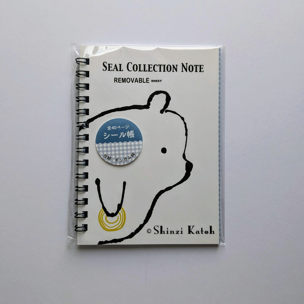 A spiral-bound notebook sealed in a clear plastic sleeve, featuring a white cover with a black bear outline, the text 'SEAL COLLECTION NOTE' and 'REMOVABLE', a blue circular sticker, and '© Shinzi Katoh' at the bottom, photographed on a light gray background.
