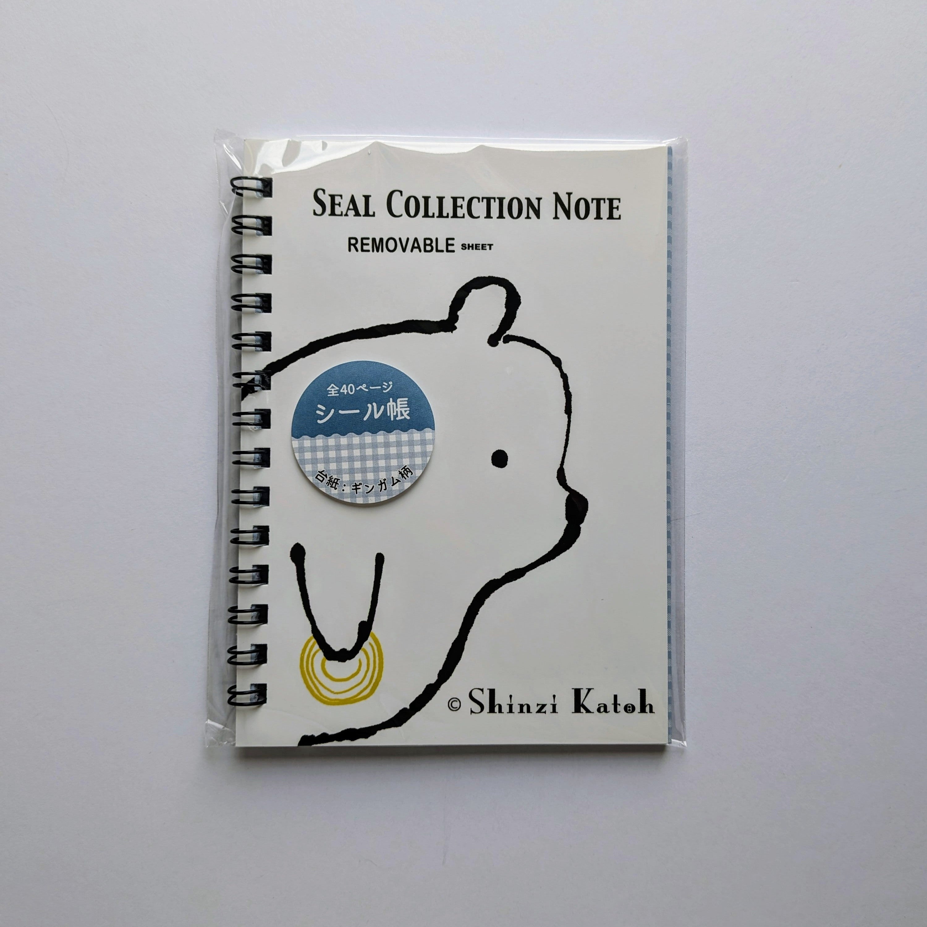 A spiral-bound notebook sealed in a clear plastic sleeve, featuring a white cover with a black bear outline, the text 'SEAL COLLECTION NOTE' and 'REMOVABLE', a blue circular sticker, and '© Shinzi Katoh' at the bottom, photographed on a light gray background.