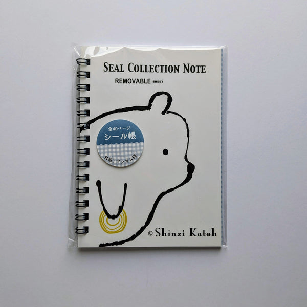 A spiral-bound notebook sealed in a clear plastic sleeve, featuring a white cover with a black bear outline, the text 'SEAL COLLECTION NOTE' and 'REMOVABLE', a blue circular sticker, and '© Shinzi Katoh' at the bottom, photographed on a light gray background.