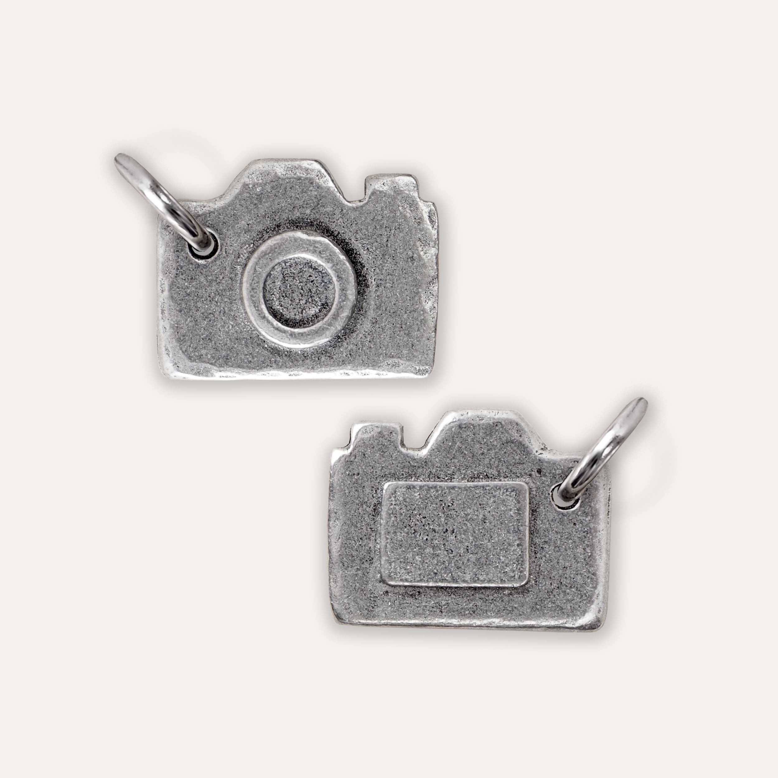 Front and reverse of silver camera-shaped charms with textured finishes and attached split rings, shown on a plain light background.