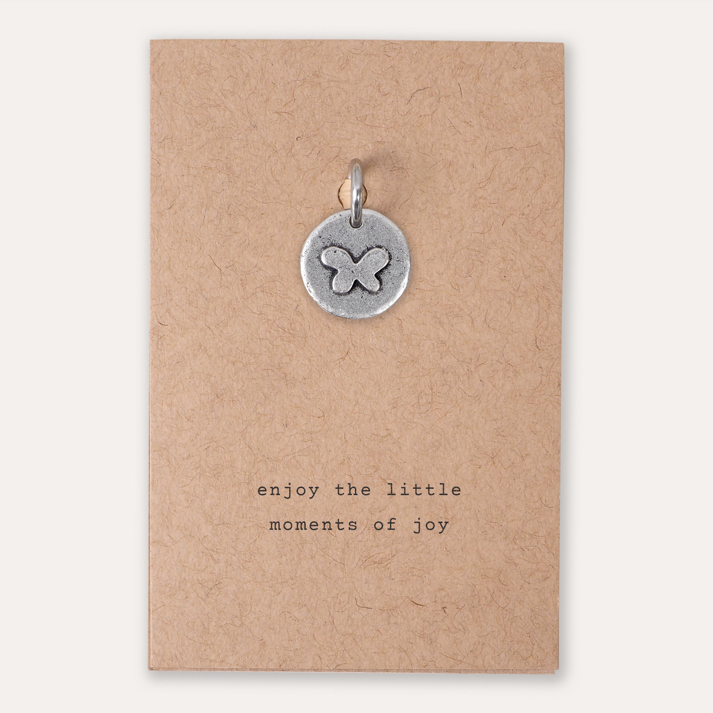 A small round silver pendant with an engraved butterfly design, displayed on a brown kraft paper card that reads 'enjoy the little moments of joy'.