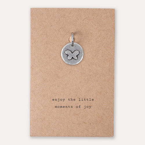 A small round silver pendant with an engraved butterfly design, displayed on a brown kraft paper card that reads 'enjoy the little moments of joy'.