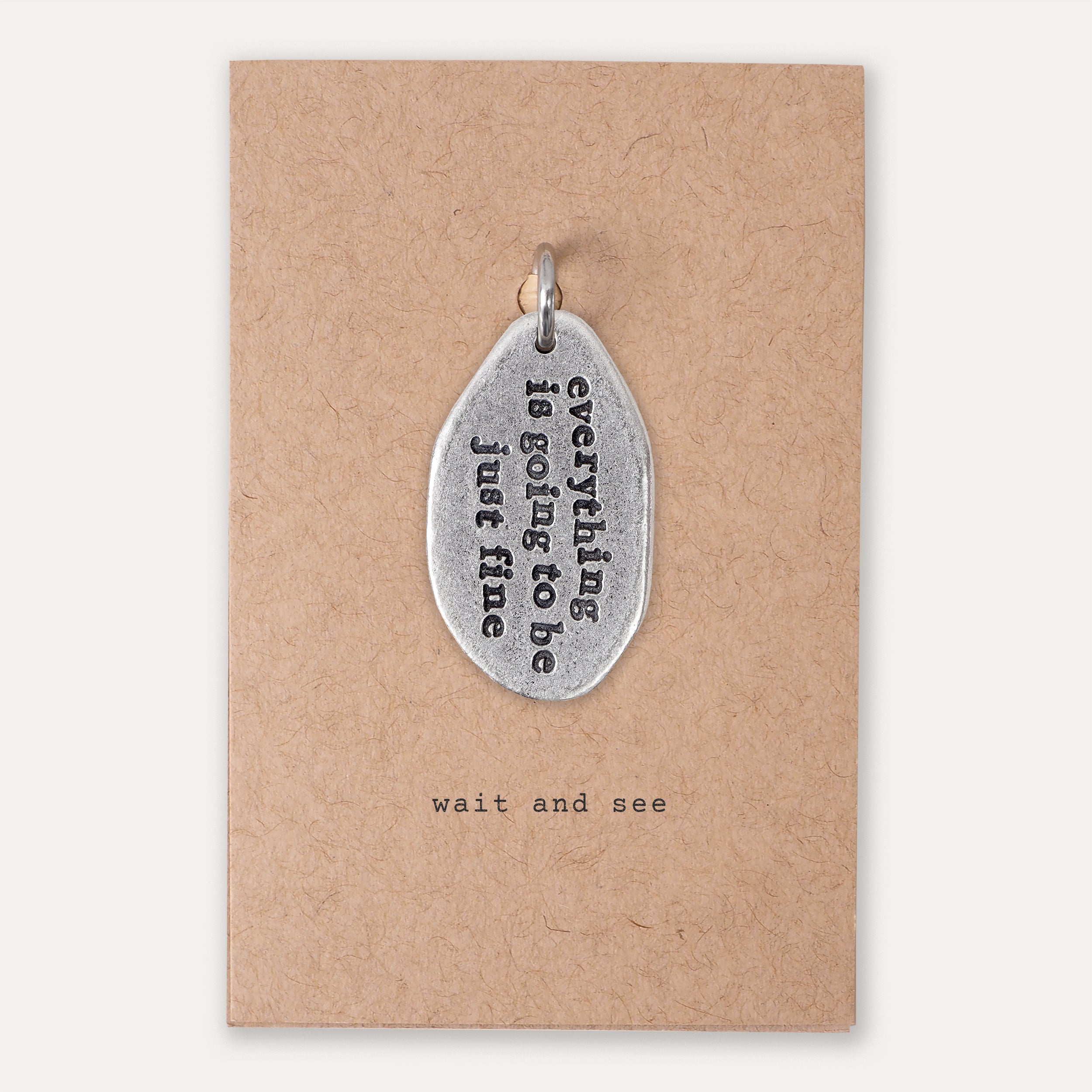 A silver-toned metal pendant with the phrase 'everything is going to be just fine' in black lettering, displayed on a brown kraft paper backing card that reads 'wait and see'.