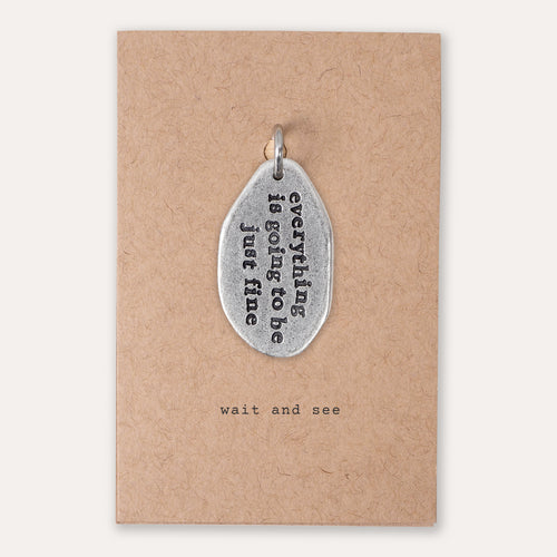 A silver-toned metal pendant with the phrase 'everything is going to be just fine' in black lettering, displayed on a brown kraft paper backing card that reads 'wait and see'.