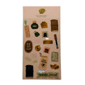 A sheet of travel-themed stickers on a light peach backing card, photographed against a plain light gray background. The stickers include motifs such as a suitcase, journal, map, passport, coffee cup, and phrases like 'ROAD TRIP', 'INK', 'LOVE TRAVEL', and 'TRAVEL journal'.